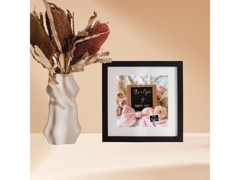 May include: A black picture frame with a white mat and a photo of a wooden sign that says "It's a Girl" with a pink bow and a sonogram photo. The photo is surrounded by pink flowers and a white fluffy background.