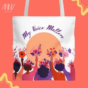 以下が含まれることがあります： 白いトートバッグ。紫色の文字で「My Voice Matters」と書かれ、桃色の太陽の上に配置されています。さまざまな色の花束を持っている多様な人物のグラフィックが特徴です。白いストラップが付いています。