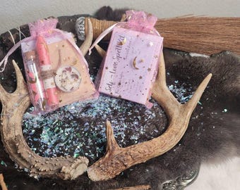 Può includere: Due piccole buste regalo rosa con un design rosa e bianco. Le buste contengono piccoli oggetti, tra cui un balsamo per le labbra, una piccola bottiglia di liquido e un piccolo oggetto rotondo. Le buste sono su una superficie marrone con corna e glitter.