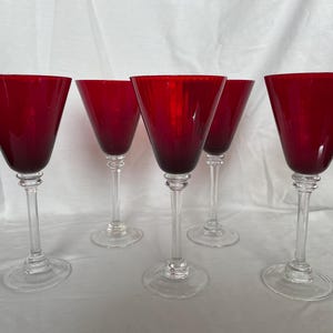 Könnte beinhalten: Fünf rote Weingläser mit klaren Stielen und Böden. Die Gläser haben eine dreieckige Schalenform. Sie sind auf einer weißen Oberfläche angeordnet. Die rote Farbe der Gläser ist lebendig.