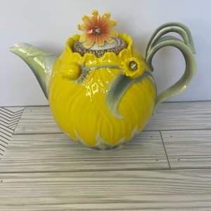 Puede incluir: Una tetera decorativa de cerámica amarilla con un diseño floral. La tetera presenta una tapa y un asa en forma de girasol, con detalles en verde y amarillo. La tetera tiene una capacidad de aproximadamente 1,5 litros.