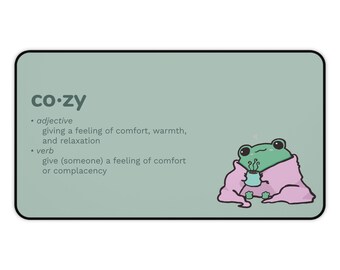 May include: A green frog wearing a pink blanket and holding a steaming cup of tea. The word "co-zy" is written in black text at the top of the image. The definition of the word "co-zy" is written in black text below the frog. The definition includes the word "adjective" and "verb".