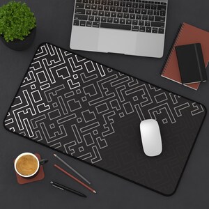 Op de afbeelding: Een zwart-witte muismat met een geometrisch patroon met een witte computermuis erop. De muismat ligt op een zwart oppervlak met een laptop, een notitieboekje en een kopje koffie.