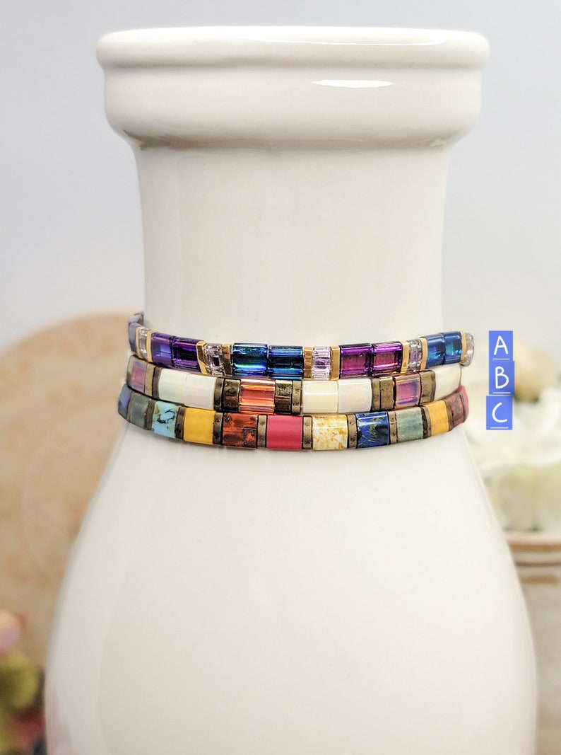 May include: Three colorful beaded bracelets stacked on a white ceramic vase. The top bracelet is purple, blue, and gold. The middle bracelet is white, orange, and gold. The bottom bracelet is yellow, red, and green.