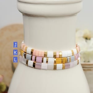 May include: Three stacked bracelets with white, pink, and gold beads. The bracelets are on a white ceramic vase.