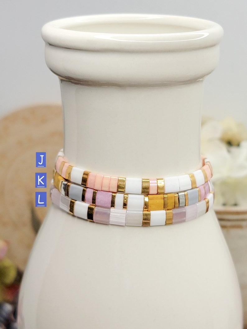 May include: Three stacked bracelets with white, pink, and gold beads. The bracelets are on a white ceramic vase.