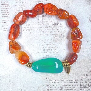 Puede incluir: Una pulsera de cuentas con piedras de cornalina naranja pulidas e irregulares dispuestas en c&iacute;rculo. Una piedra verde &uacute;nica, lisa y alargada, se sit&uacute;a en la parte inferior, flanqueada por dos peque&ntilde;os espaciadores met&aacute;licos de tono dorado.