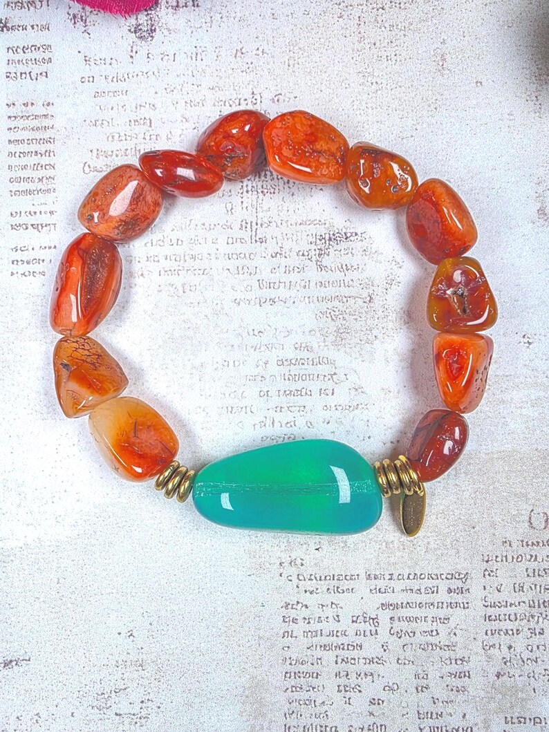 Puede incluir: Una pulsera de cuentas con piedras de cornalina naranja pulidas e irregulares dispuestas en c&iacute;rculo. Una piedra verde &uacute;nica, lisa y alargada, se sit&uacute;a en la parte inferior, flanqueada por dos peque&ntilde;os espaciadores met&aacute;licos de tono dorado.