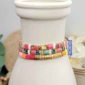 May include: Three colorful beaded bracelets stacked on a white ceramic vase. The bracelets are made of small, square beads in shades of pink, orange, yellow, blue, green, and white. The top bracelet has gold beads interspersed throughout.