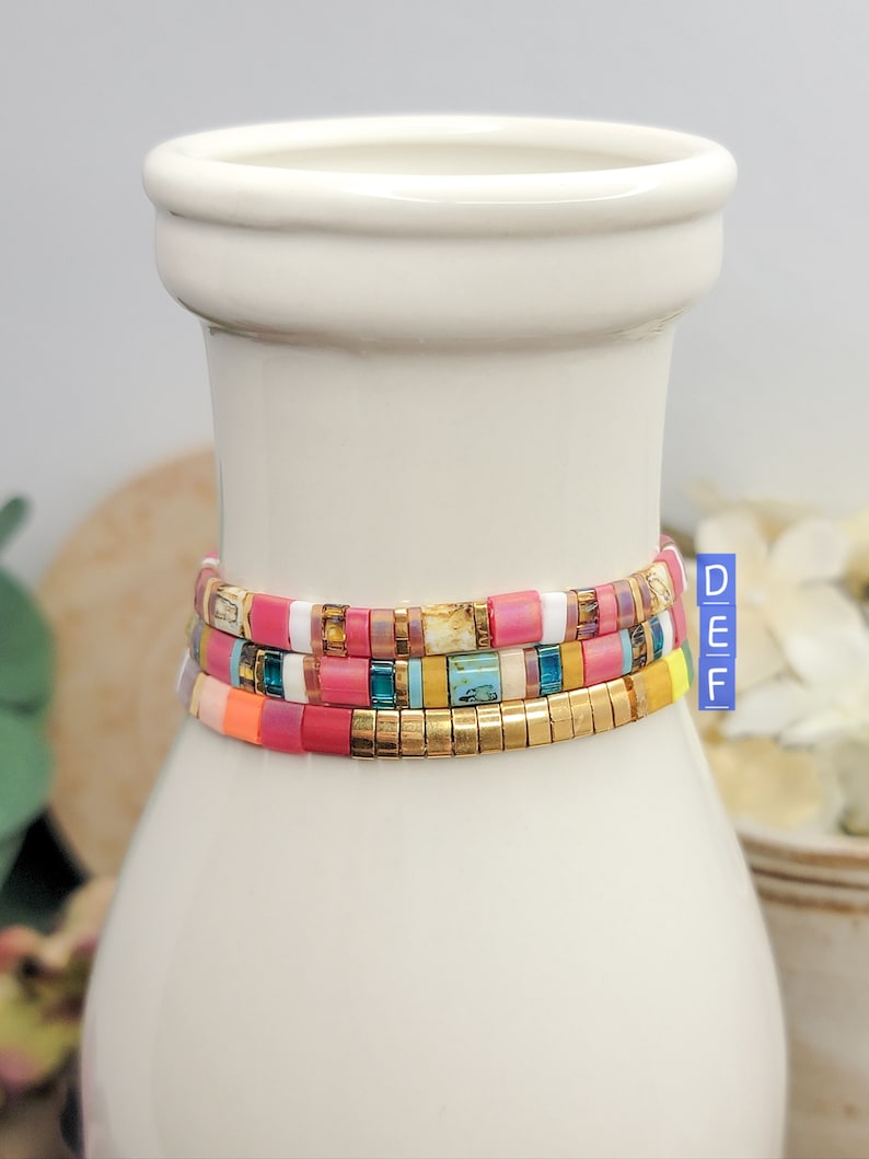 May include: Three colorful beaded bracelets stacked on a white ceramic vase. The bracelets are made of small, square beads in shades of pink, orange, yellow, blue, green, and white. The top bracelet has gold beads interspersed throughout.