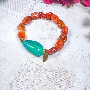 Puede incluir: Una pulsera el&aacute;stica con cuentas de cornalina naranja pulidas e irregulares y una cuenta grande, lisa y con forma de l&aacute;grima de color verde azulado. La cuenta verde azulado est&aacute; flanqueada por dos espaciadores de metal dorado y un dije peque&ntilde;o.