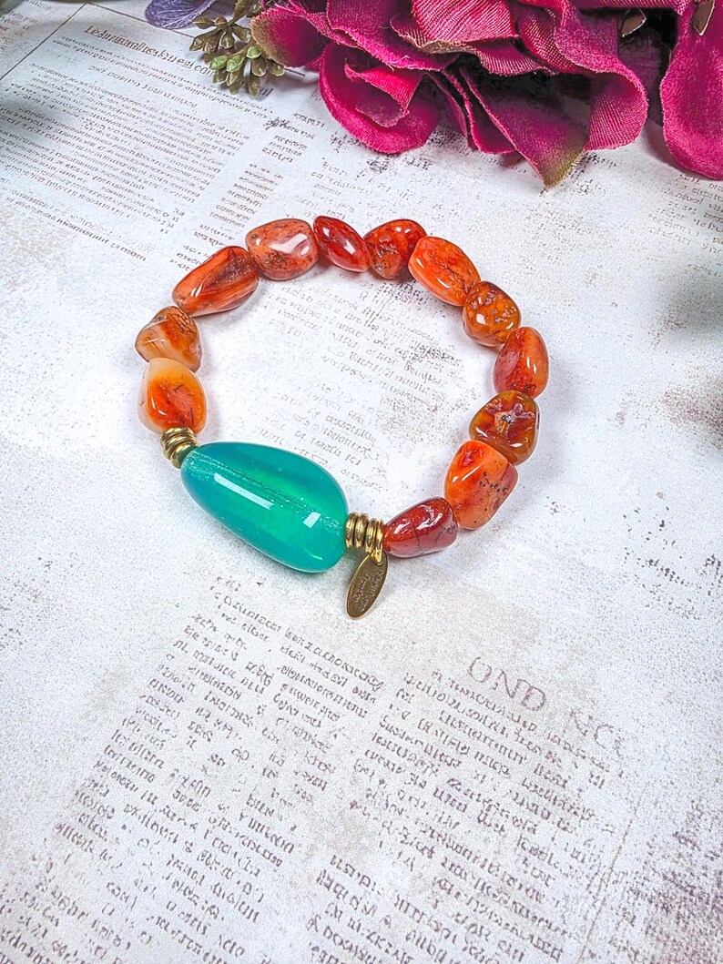 Puede incluir: Una pulsera el&aacute;stica con cuentas de cornalina naranja pulidas e irregulares y una cuenta grande, lisa y con forma de l&aacute;grima de color verde azulado. La cuenta verde azulado est&aacute; flanqueada por dos espaciadores de metal dorado y un dije peque&ntilde;o.