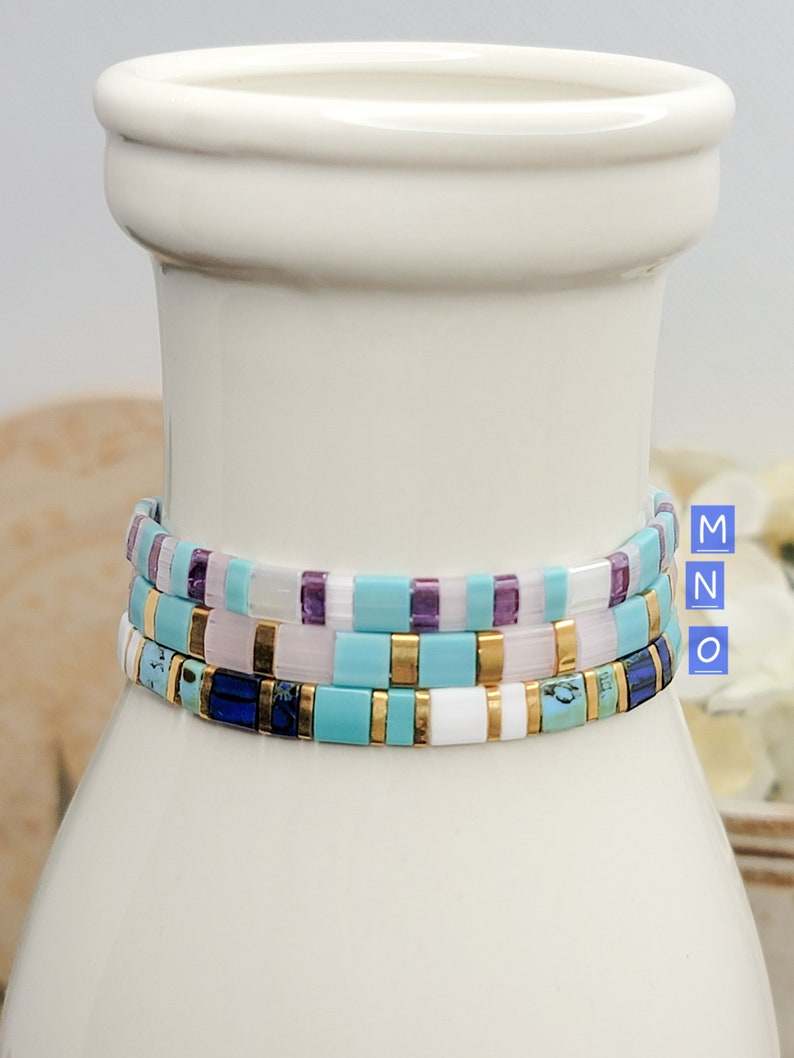 May include: Three stacked bracelets with turquoise, white, and gold beads. The top bracelet has purple beads, the middle bracelet has pink beads, and the bottom bracelet has blue beads.
