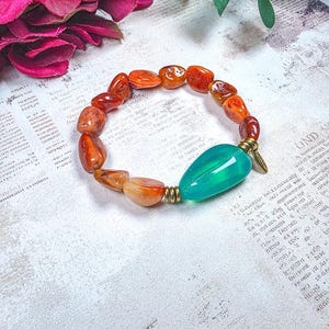 Puede incluir: Una pulsera el&aacute;stica con cuentas de cornalina naranja pulidas e irregulares y una cuenta &uacute;nica, lisa y en forma de l&aacute;grima de color verde azulado. La cuenta verde azulado tiene espaciadores met&aacute;licos dorados antiguos y un peque&ntilde;o dije colgante.