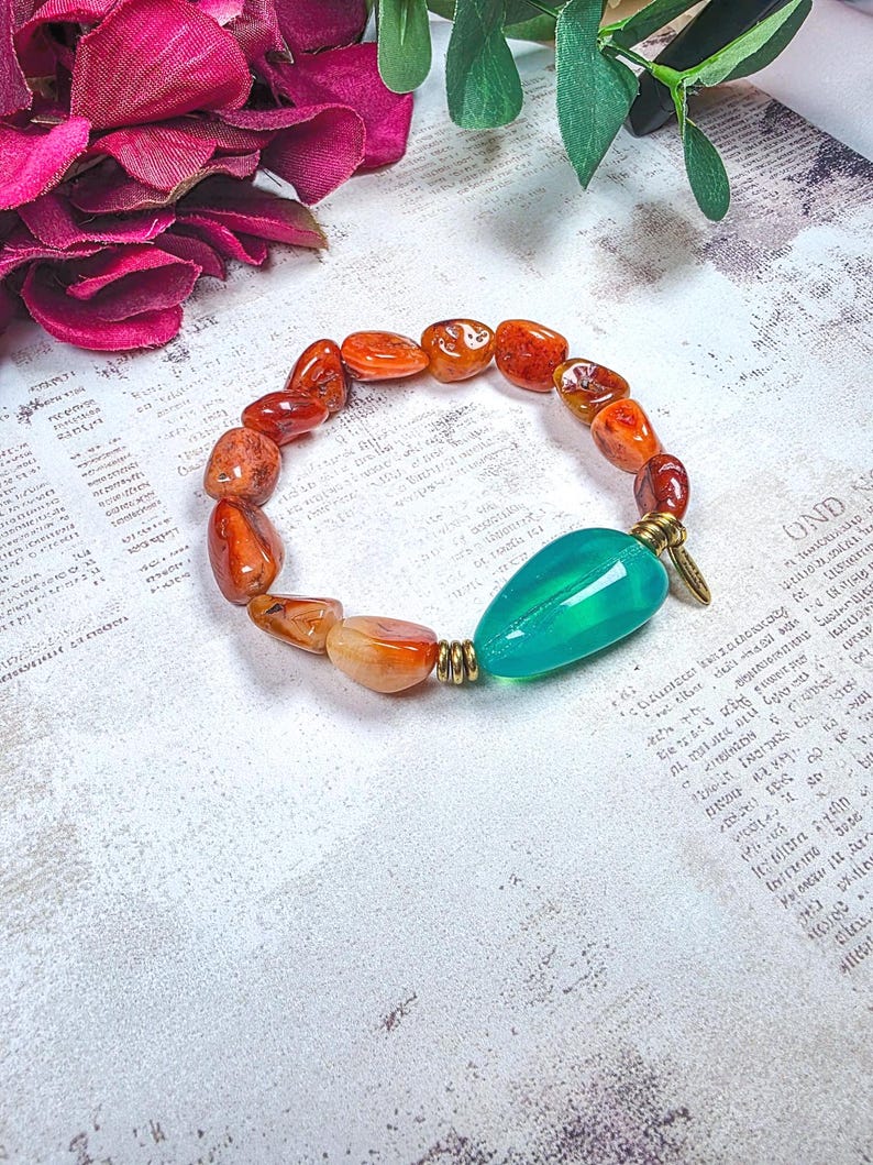 Puede incluir: Una pulsera el&aacute;stica con cuentas de cornalina naranja pulidas e irregulares y una cuenta &uacute;nica, lisa y en forma de l&aacute;grima de color verde azulado. La cuenta verde azulado tiene espaciadores met&aacute;licos dorados antiguos y un peque&ntilde;o dije colgante.
