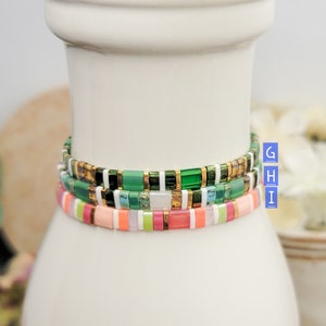 May include: Three stacked bracelets with colorful square beads. The top bracelet has green, gold, and white beads. The middle bracelet has green, white, and pink beads. The bottom bracelet has pink, green, and white beads.