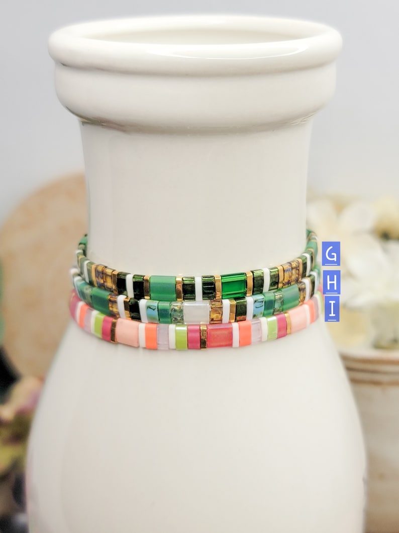May include: Three stacked bracelets with colorful square beads. The top bracelet has green, gold, and white beads. The middle bracelet has green, white, and pink beads. The bottom bracelet has pink, green, and white beads.