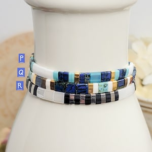 May include: Three stacked bracelets with colorful square beads. The top bracelet is white with gold accents, the middle bracelet is blue and turquoise with gold accents, and the bottom bracelet is black and white with silver accents.