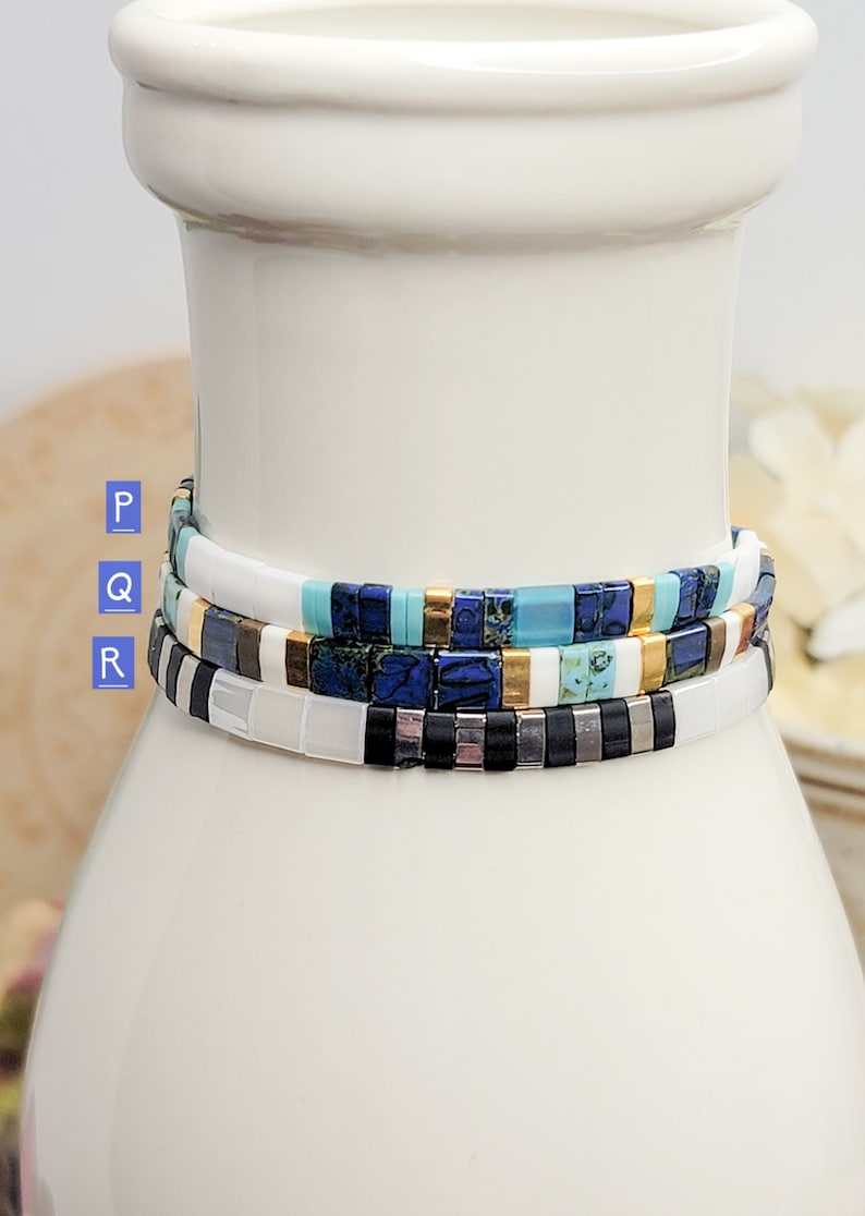 May include: Three stacked bracelets with colorful square beads. The top bracelet is white with gold accents, the middle bracelet is blue and turquoise with gold accents, and the bottom bracelet is black and white with silver accents.