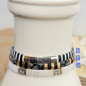 May include: Three stacked bracelets with black and white striped, black and gold, and white beads. The bracelets are on a white ceramic vase.