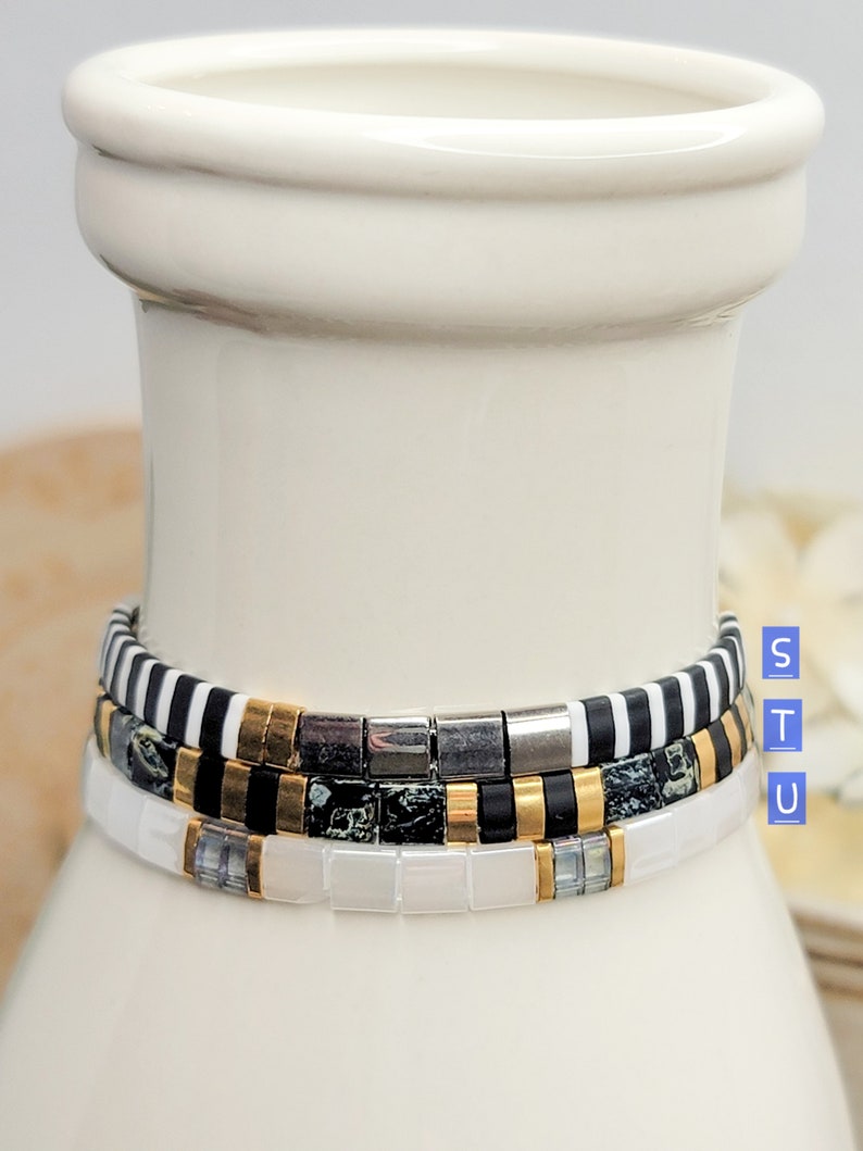 May include: Three stacked bracelets with black and white striped, black and gold, and white beads. The bracelets are on a white ceramic vase.