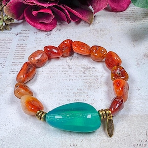 Puede incluir: Una pulsera el&aacute;stica con cuentas de cornalina naranja pulidas e irregulares y una sola cuenta de vidrio grande, lisa y transl&uacute;cida de color verde azulado. Incluye dos espaciadores de metal dorado y un peque&ntilde;o dije ovalado de metal dorado.