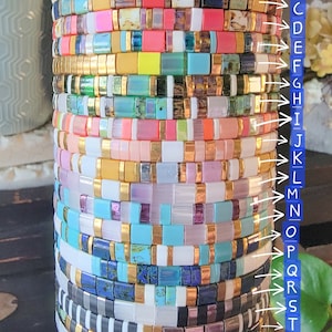 May include: A stack of 26 colorful beaded bracelets with gold accents. Each bracelet is made of small square beads in various colors and patterns.