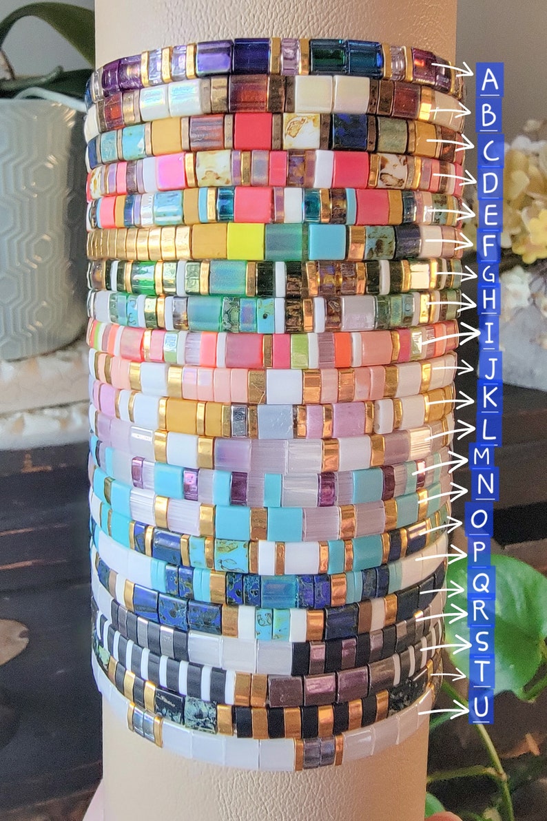 May include: A stack of 26 colorful beaded bracelets with gold accents. Each bracelet is made of small square beads in various colors and patterns.