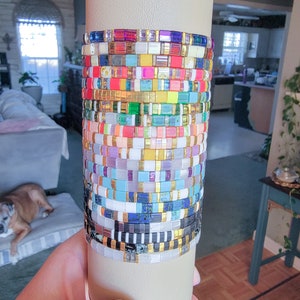 May include: A stack of colorful and patterned bracelets made of small square tiles. The bracelets are arranged in a vertical stack on a white background.