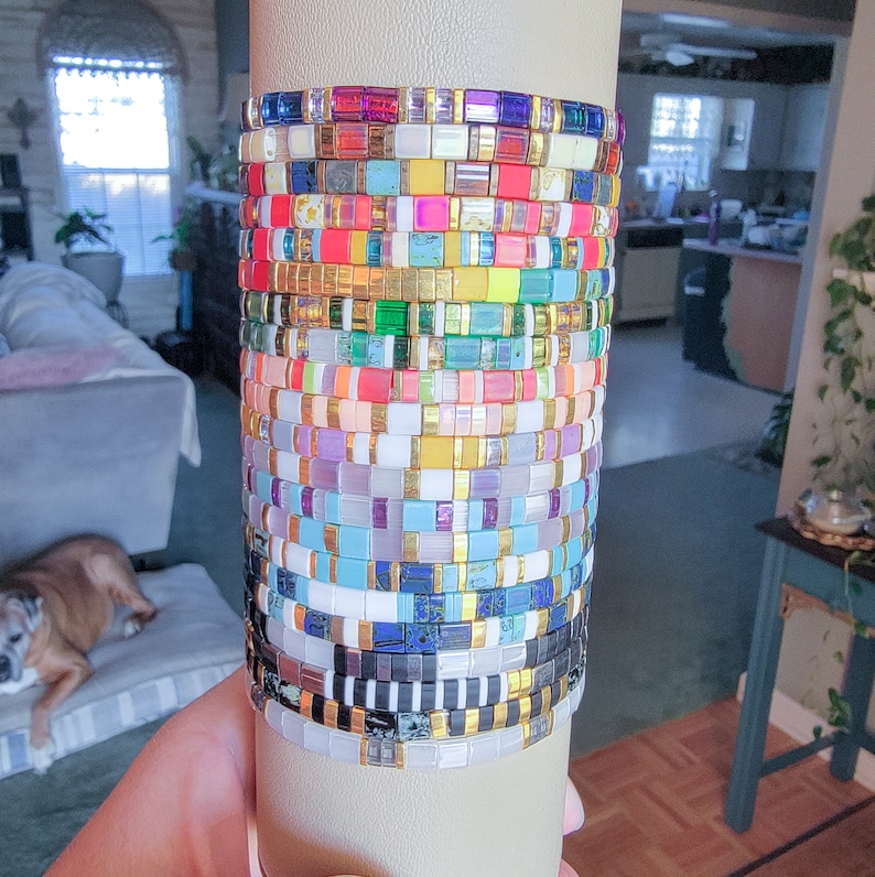May include: A stack of colorful and patterned bracelets made of small square tiles. The bracelets are arranged in a vertical stack on a white background.
