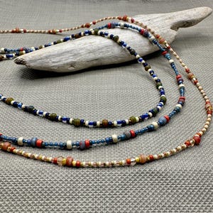 May include: Three layered necklaces featuring a variety of small beads in shades of blue, red, green, tan, and clear glass. The strands are arranged in a cascading pattern, showcasing a mix of matte, metallic, and translucent bead textures.