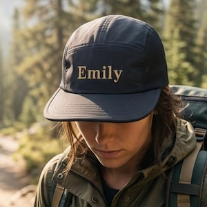 May include: A navy blue cap with the name "Emily" embroidered in gold lettering. The cap has a flat brim and is worn by a person wearing a green jacket and carrying a backpack. The background shows a forest trail.