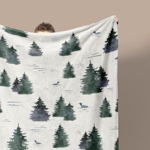 May include: A white blanket with a repeating pattern of watercolor-style evergreen trees and small duck illustrations in shades of green and gray. The blanket is held up by a person.