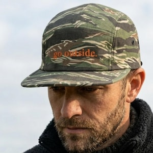 May include: A green and brown camouflage cap with the words "go outside" in orange. The cap has a flat peak and eyelets. The hat is made of a durable fabric and is suitable for outdoor activities. The cap is a stylish accessory.