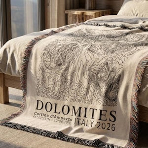 May include: Beige throw blanket with a black topographical map design and fringed edges. The text "DOLOMITES" and "ITALY 2026" are printed on the blanket, along with coordinates. The blanket is displayed on a bed.
