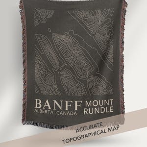 May include: Dark gray throw blanket featuring a topographical map of Banff and Mount Rundle in Alberta, Canada. The map is in white, with the text "Banff Mount Rundle" and "Accurate Topographical Map". The blanket has a fringed edge.