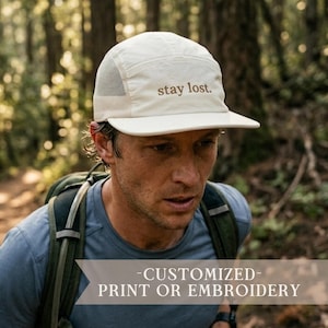 May include: A cream-colored five-panel cap featuring a flat brim and mesh side panels. The front of the cap displays the text "stay lost." embroidered in a brown font. The hat is constructed with a structured crown and an adjustable back strap.