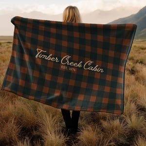 May include: A person holding a large, rectangular blanket with a brown and dark green plaid pattern. The blanket has the text "Timber Creek Cabin EST. 1974" in a cursive font.