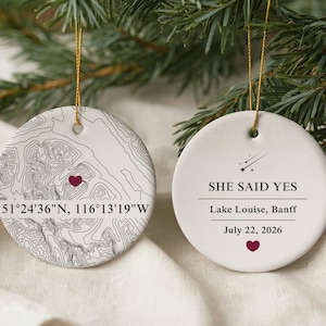 May include: Two round white ceramic ornaments with gold hanging strings. One features a black topographic map with a maroon heart and coordinates 51°24'36"N, 116°13'19"W. The other displays a shooting star icon, the text SHE SAID YES, Lake Louise, Banff, July 22, 2026, and a maroon heart.