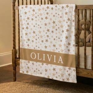 May include: A white baby blanket with a pattern of gold stars. The blanket has a gold band with the name "OLIVIA" in white letters. The blanket is draped over a wooden crib.