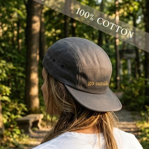 May include: A dark grey, five-panel cap made of 100% cotton. The cap has a flat peak and the words "go outside" are embroidered in gold. The background is a blurred forest scene.