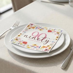 May include: A white plate setting with a floral-decorated napkin that reads "90 years loved." The napkin is surrounded by colorful flowers. A fork and knife are placed on either side of the plate. The table is covered with a light beige tablecloth.