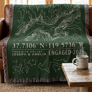 May include: A dark green woven throw blanket featuring white topographic map lines, the coordinates 37.7306 N 119.5736 W, the text Yosemite Valley, Joseph & Amelia, and Engaged 2026. The blanket has a fringed edge and a textured fabric construction.