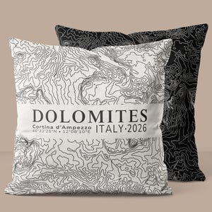 May include: Two cushions with a topographical map design. One cushion is white with a black map design and the word "DOLOMITES" printed on it. The other cushion is black with a white map design. The text "Cortina d'Ampezzo ITALY 2026" is also printed on the white cushion.