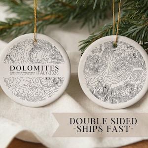 May include: Two white, round Christmas ornaments with a topographic map design. One ornament features the word "DOLOMITES" and "ITALY-2026". Both ornaments have a gold string for hanging. The ornaments are displayed on a white cloth with a green pine branch in the background.