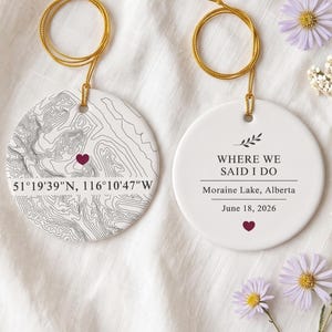 May include: Two round white ceramic ornaments with gold hanging strings. One features a black topographic map with a small maroon heart and coordinates 51°19'39"N, 116°10'47"W. The other displays the text WHERE WE SAID I DO, Moraine Lake, Alberta, June 18, 2026, and a maroon heart.