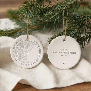 May include: Two round white ceramic ornaments with gold hanging strings. One ornament features a topographic map, coordinates 39°10'20.0"N 120°08'20.0"W, and the text WHERE ONLY THE BEST MEMORIES ARE MADE. The other displays THE SMITH CABIN EST. 2026.