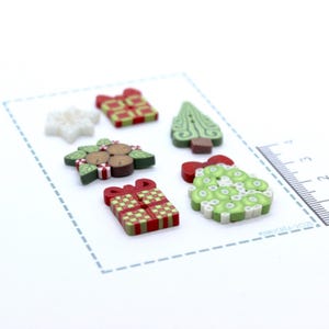 Pode incluir: Uma coleção de botões coloridos com tema natalino. Os botões incluem uma estrela branca, uma árvore de Natal verde, uma coroa verde e caixas de presente com laços vermelhos. Os botões estão em uma superfície branca.