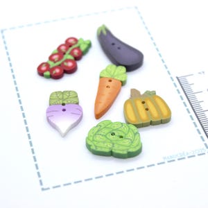 Puede incluir: Un conjunto de botones coloridos con forma de verduras. El conjunto incluye un racimo de tomates rojos, una berenjena morada, una zanahoria naranja, una calabaza amarilla, un nabo morado y una col verde. Cada botón tiene un ojal.