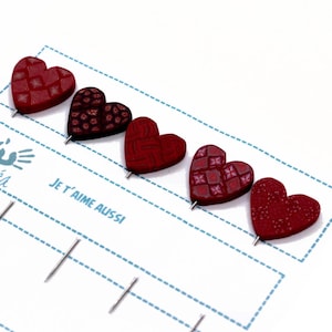 Könnte beinhalten: Fünf rote herzförmige Stecknadeln mit verschiedenen Mustern. Die Stecknadeln sind in einer Reihe auf einer weißen Karte mit blauen gestrichelten Linien und dem Text "Je t'aime aussi" angeordnet.